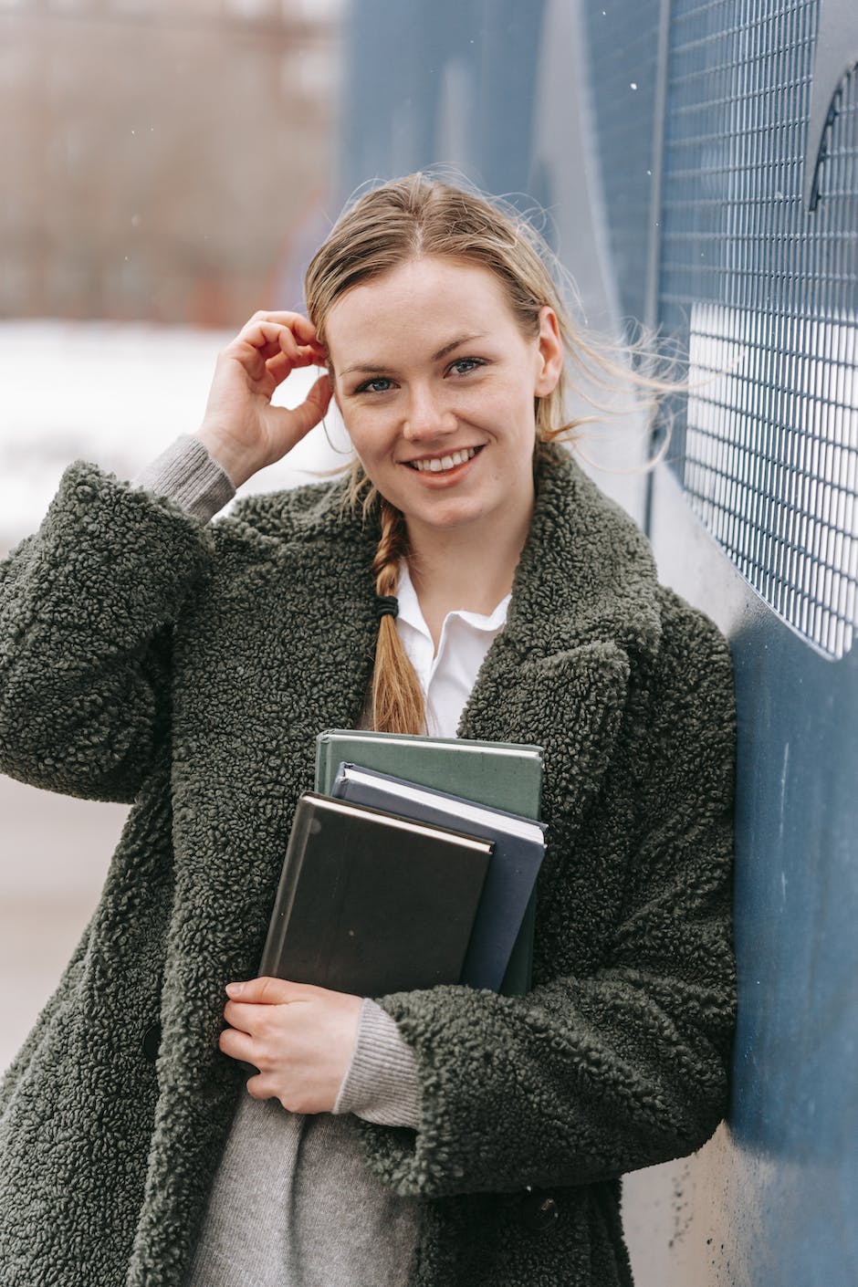 Smiling young lady with textbooks looking at camera against the wall in town on a windy day